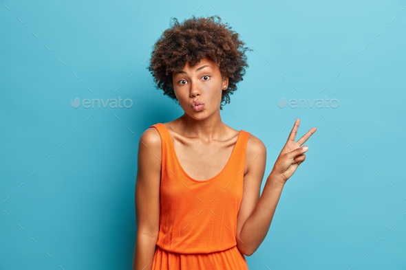 Pleasant looking young Afro American woman keeps lips folded and makes peace gesture has good mood e