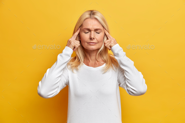 Serious elderly wrinkled woman keeps fingers on temples and closes eyes to relieve severe headache t - Stock Photo - Images