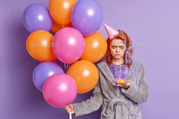 Unhappy angry redhead birthday girl smeared with serpentine spray has leaked makeup holds cupcake ba