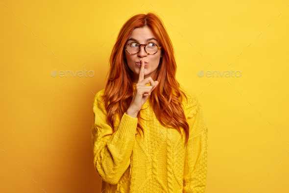 Red haired woman holds index finger on lips makes quiet gesture tells secret information shares some