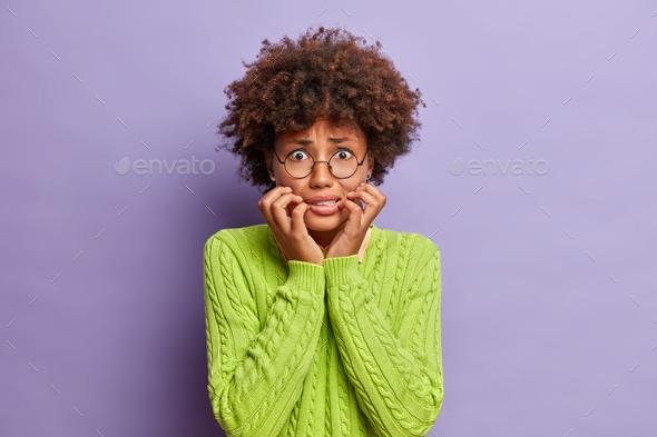 Photo of dark skinned worried woman bites finger nails and looks anxious at camera worries before im