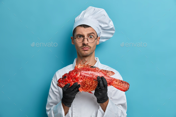Photo of serious male cook carries fish looks directly at camera asks chef for advice what better to