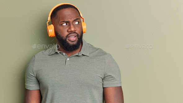 Portrait Of Serious Handsome Caucasian Male Listens Music Or Audio Book  Uses Modern Wireless Headphones Poses Against Grey Background With Copy  Space For Your Information Advertising Or Text Stock Photo - Download