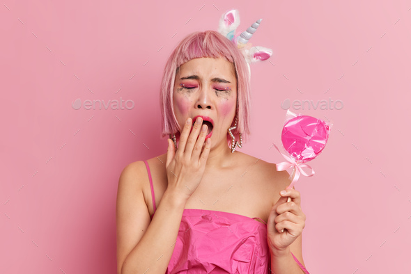 Unhappy dissatisfied young woman with pink hair covers mouth as wants to sleep holds delicious candy