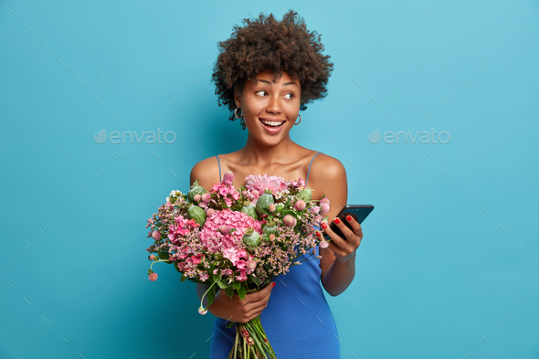 Positive dark skinned woman with curly hair holds nice bouquet of flowers gets order online uses mod