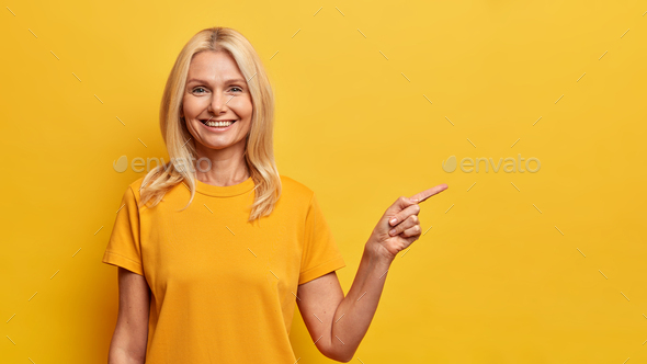 Portrait of pleasant looking blonde woman with satisfied expression points at blank space dressed in