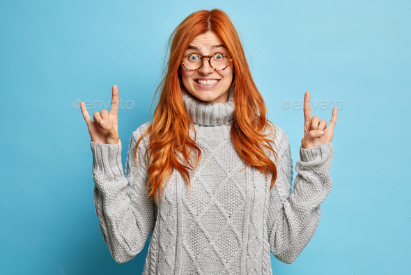 Pleased joyous redhead woman in spectacles shows rock n roll hand sign devil horn gesture smiles bro