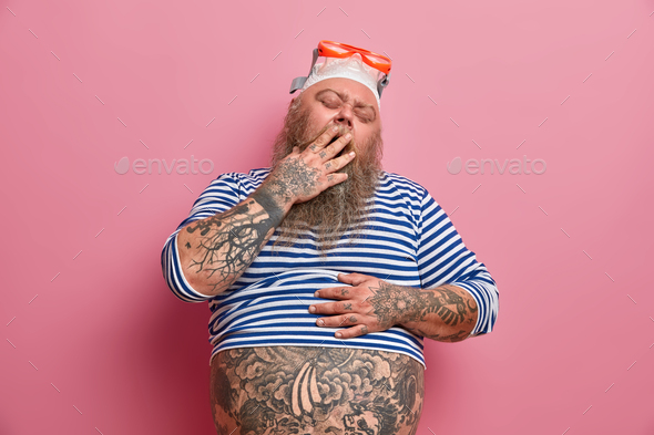 Tired fatso man being lazy wants to sleep yawns and covers mouth recreats at sea wears swim hat gogg