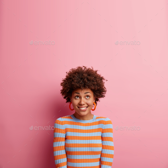 Vertical shot of happy curious woman with Afro hair bites lips and looks upwards dressed in casual j