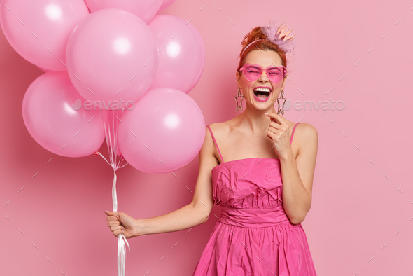 Cheerful redhead woman dressed in pink dress and sunglasses has fun on graduation party poses with b