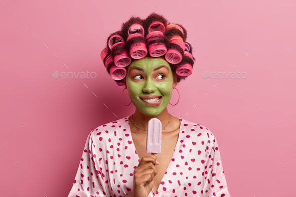 Pleased young African American woman bites lips applies green beauty mask wears hair curlers eats de