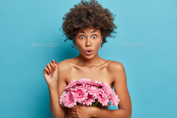 Shocked curly haired woman with naked body holds breath from wonder carries pink gerbera flowers rai