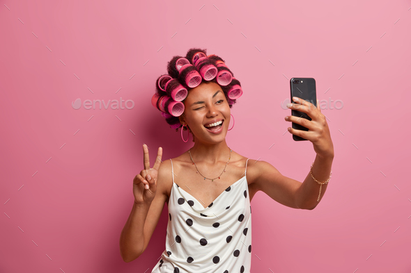 Photo of cheerful dark skinned woman applies hair curlers winks eye and makes peace gesture poses in