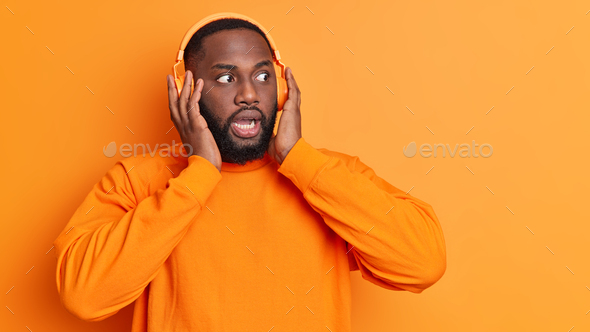 Dark skinned bearded man stares bugged eyes keeps hands on stereo headphones listens music reacts on