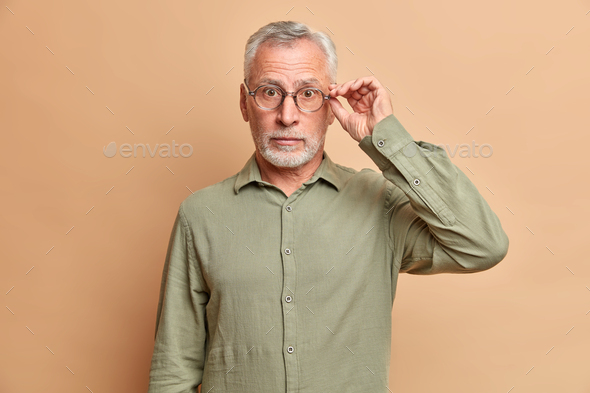 Surprised grey haired handsome man keeps hand on rim of spectacles hears amazing news wears formal s