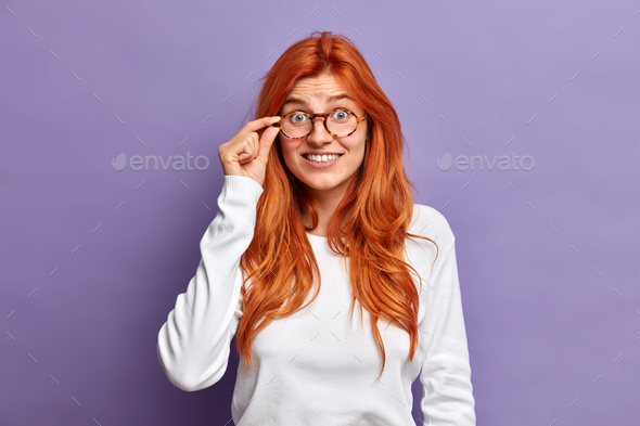 Waist up shot of lovely redhead woman keeps hand on rim of spectacles looks with interest hears some