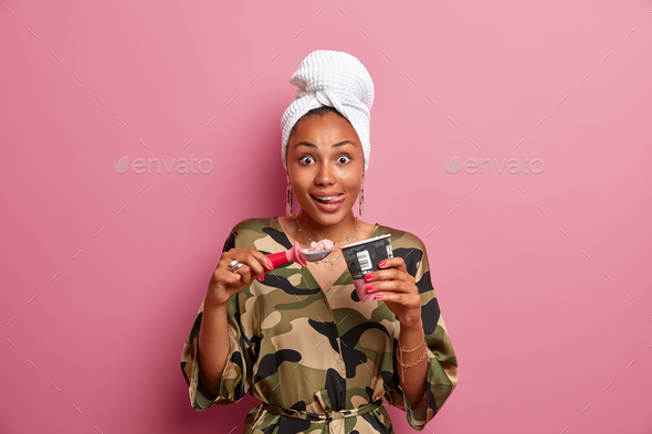 Photo of beautiful surprised woman with dark skin licks lips with tongue as eats delicious ice cream