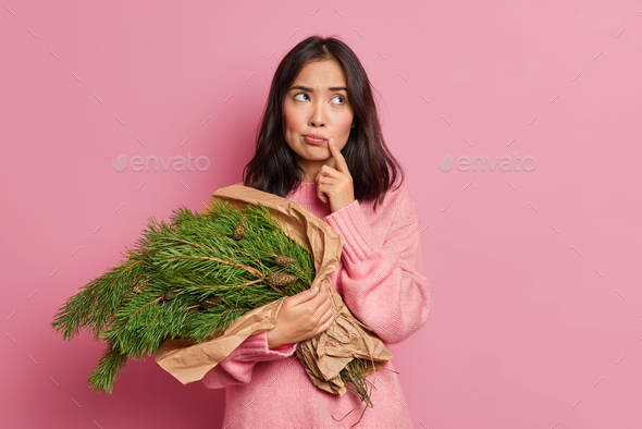 Horizontal shot of serious Asian woman makes plans in mind keeps finger near lips holds fir tree bou