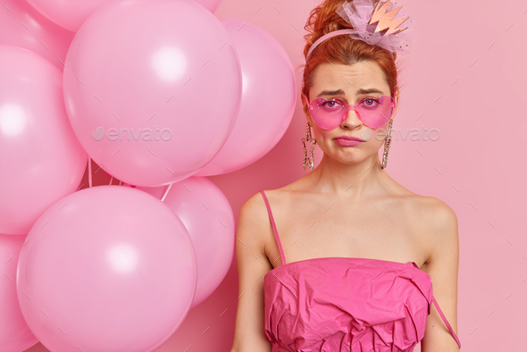 Displeased young female with professional makeup wears pink sunglasses and dress looks unhappily at