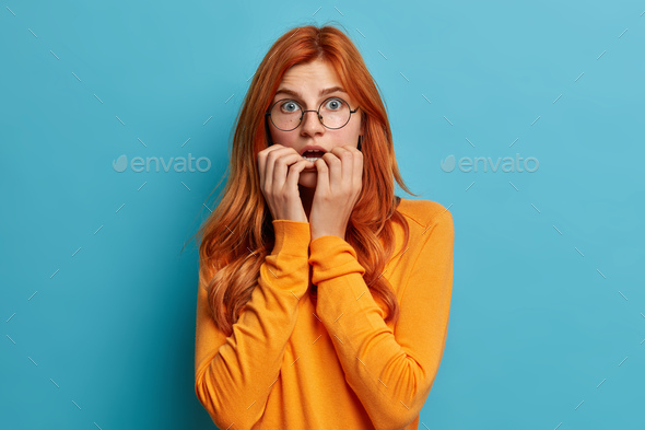 Puzzled surprised redhead Caucasian woman bites finger nails keeps mouth opened gasps from amazement