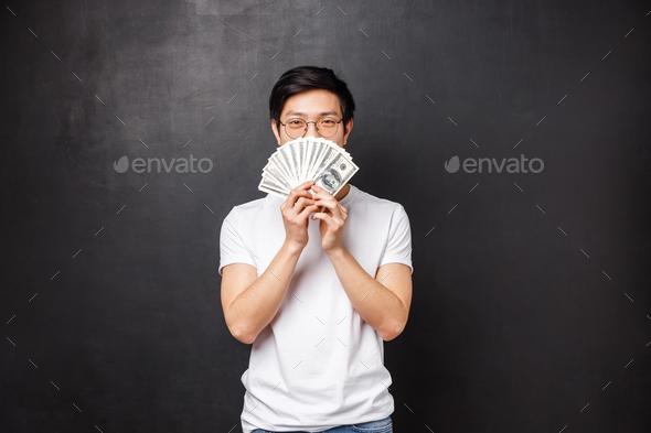 Portrait of sly happy and pleased asian young man winning big cash prize money, hiding face behind