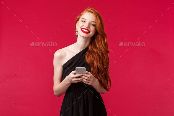 Portrait of gorgeous smiling woman with ginger hair in elegant black prom dress, holding smartphone
