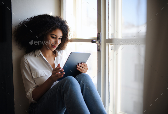 Portrait of young woman on window sill indoors at home, using tablet.