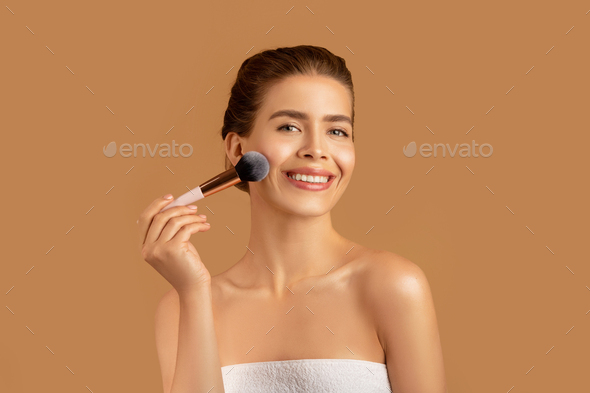Pretty young lady using cosmetic brush to apply decorative makeup on her face, brown studio