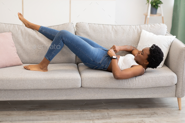 Black Woman Struggling Zipping Small Jeans Lying On Sofa Indoors