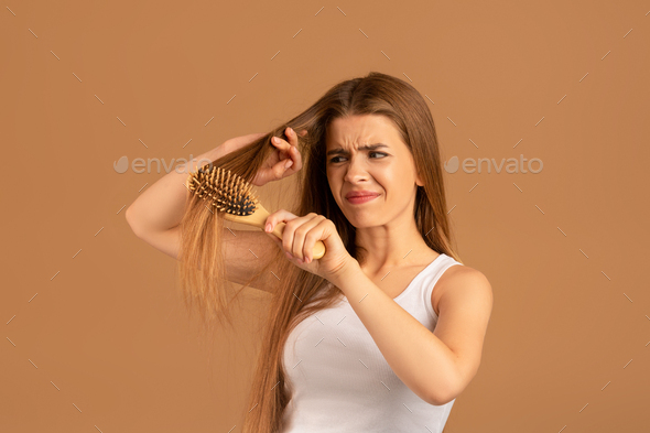 Annoyed young woman having problem brushing her long hair on brown studio background. Hairdressing