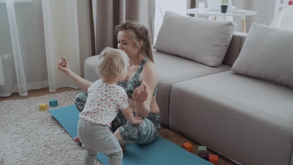 Happy Mindful Mother Doing Morning Exercises in Yoga Pose While Kid Daughter Playing at Home alt