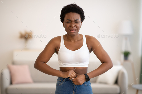 Frustrated Black Woman Buttoning Small Jeans After Weight Gain Indoors