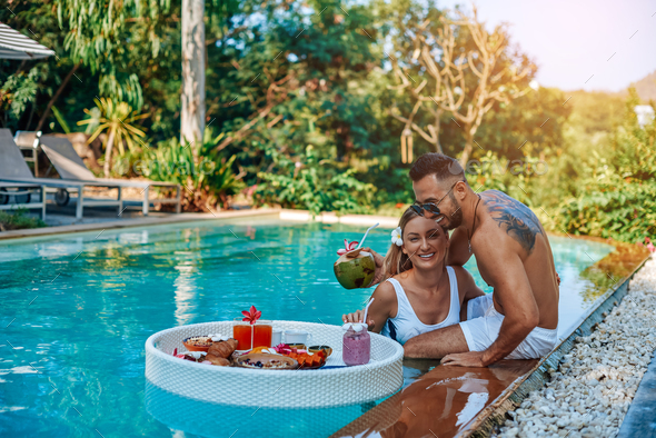 Laughing couple of travellers swim in a pool with floating table with food