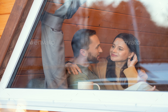 Young serene affectionate couple interacting by large window of country house