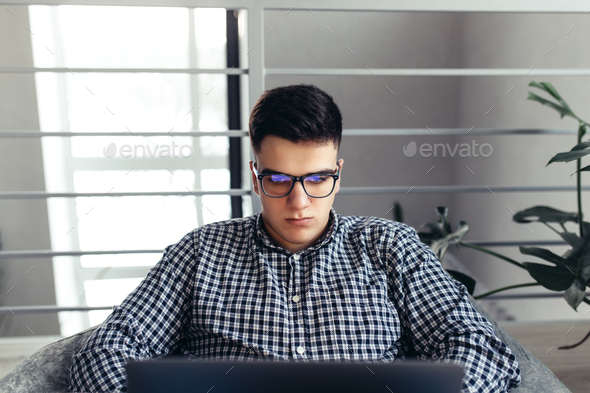 Man using laptop, wearing glasses, cushioned furniture. Blurred background