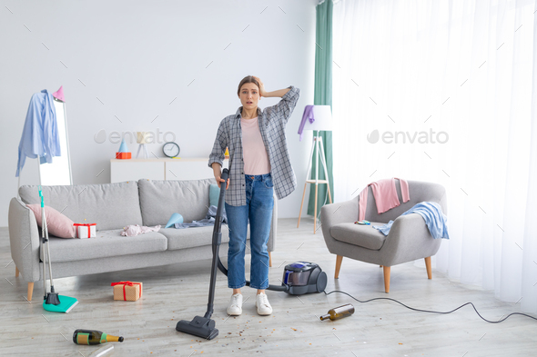 After party chaos. Dismayed millennial woman standing in her messy apartment with vacuum cleaner