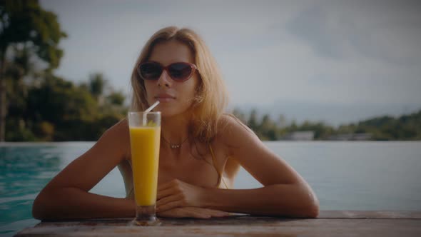 A Blonde Woman Drinks Mango Shake While Lounging in the Sun By the Pool on Vacation alt