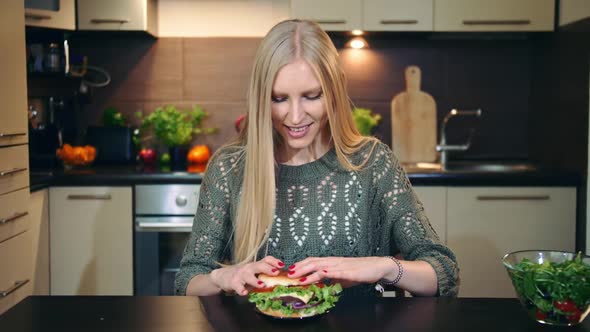 Young Woman Eating Fast Food, Hamburger While Sitting at Table in Stylish Kitchen. alt