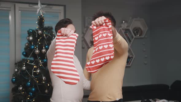 Joyful Gay Couple in Love Holding Christmas Stockings in Front of the Christmas Tree alt