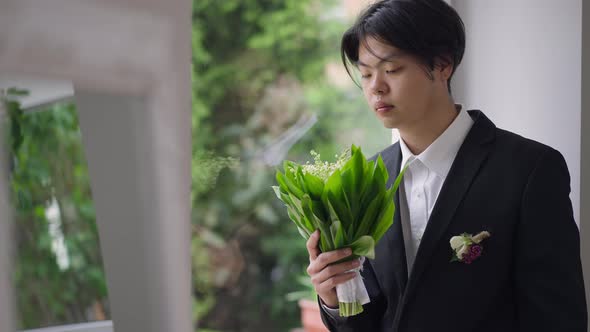 Romantic Confident Asian Groom Admiring Bouquet of Lilies of the Valley and Looking Out the Window alt