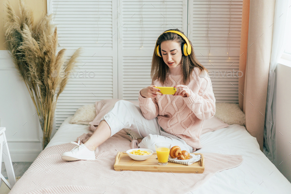 Millennial girl makes a photo of breakfast on a tray listening to music on headphones.
