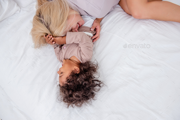 Young Caucasian blond mother tickles little African American daughter. Lie on white bed, having fun