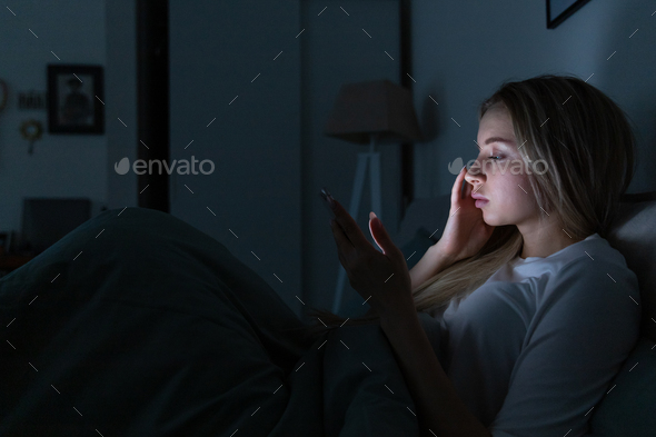 Sleepy exhausted woman lying in bed using cellphone chatting in social networks, bored can not sleep