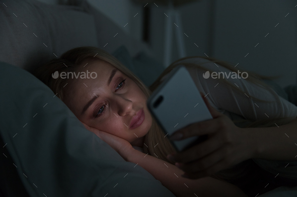 Young sleepy woman lying in bed under the blanket using smartphone at night, can not sleep. Insomnia