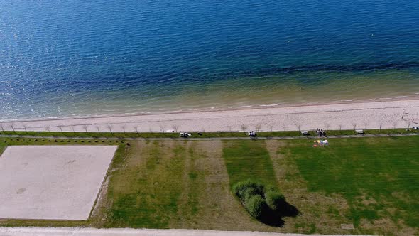 Lake beach with people sunbathing and bathing, garden and walking area. Overhead drone shot travelin alt