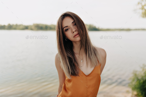 Outside portrait of adorable woman with dark hair wearing orange blouse on the background of lake