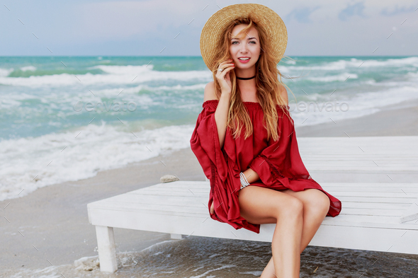 Ginger pretty woman in stylish straw hat and red beach dress sitting near  the ocean. Stock Photo by ssvetasokolova