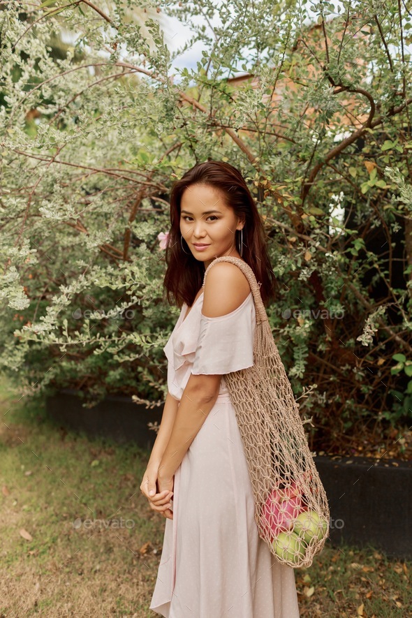 Asian woman in dress holding eco friendly mesh shopper bag with fresh tropical fruits.