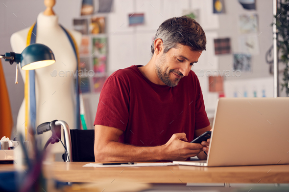 Mature Male Fashion Designer In Wheelchair Using Mobile Phone To Browse Internet Sitting At Desk
