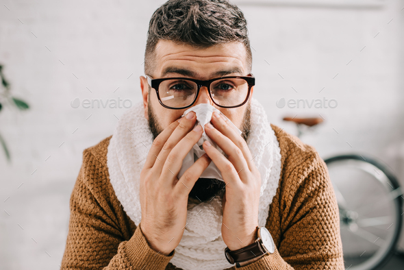 portrait of sick man in knitted scarf sitting in office, sneezing and covering mouth with tissue